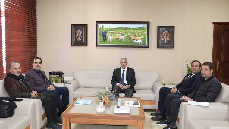 Bir odada beş kişi koltuklarda oturuyor; ortadaki kişinin önünde sehpa ve duvarında koyun tablosu bulunuyor.