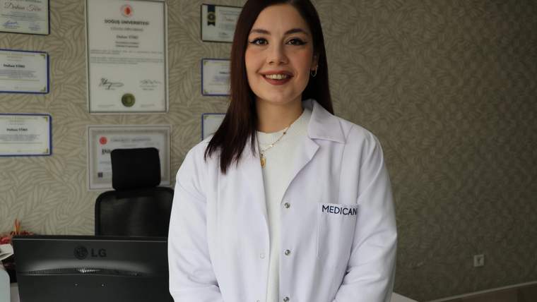 Beyaz lab coat giymiş bir kadın, ofis ortamında gülümsüyor.
