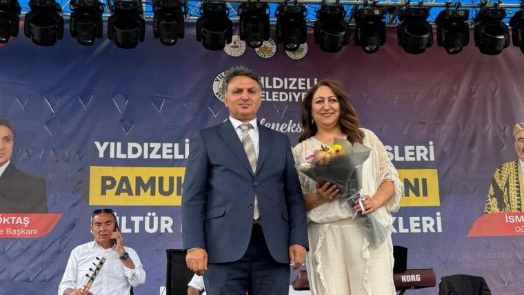 Bir erkek ve bir kadın, sahnede çiçekle birlikte poz veriyor. Arkada pankartta "Pamukpınar Kültür Festivali" yazıyor.