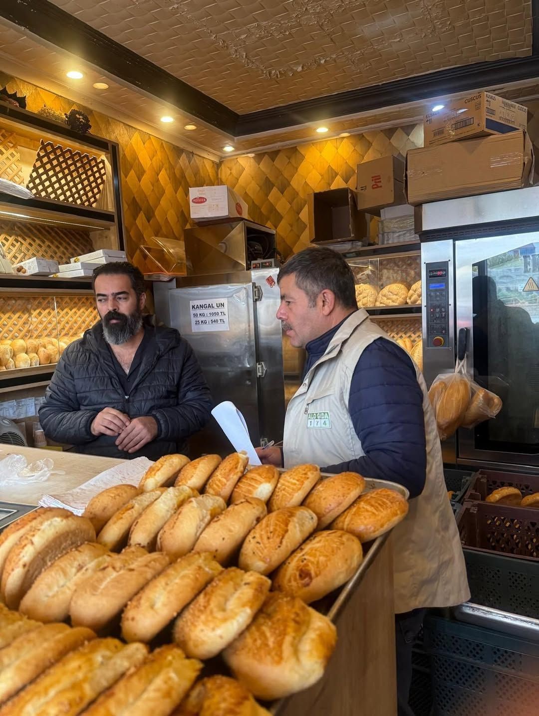 Ekmeklerle dolu tezgâhın arkasında iki erkek, fırının içinde yan yana duruyor.