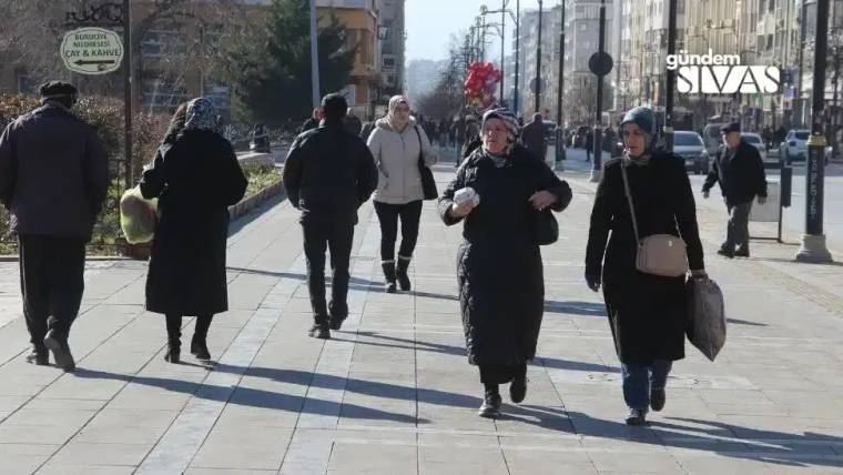 Geniş bir kaldırımda yürüyen, çoğu koyu renk giyinmiş çok sayıda kişi görülüyor.