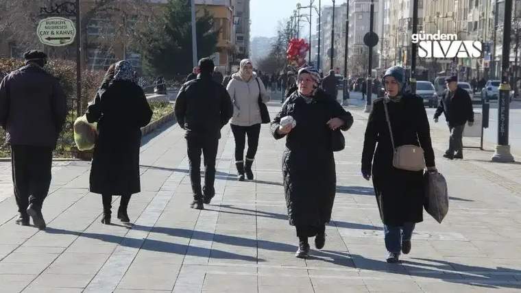Geniş bir kaldırımda yürüyen, çoğu koyu renk giyinmiş çok sayıda kişi görülüyor.