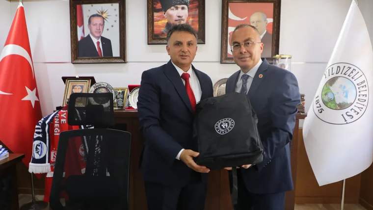 İki kişi, masada bir çanta tutarak poz veriyor; arka planda Türk bayrağı ve portreler var.