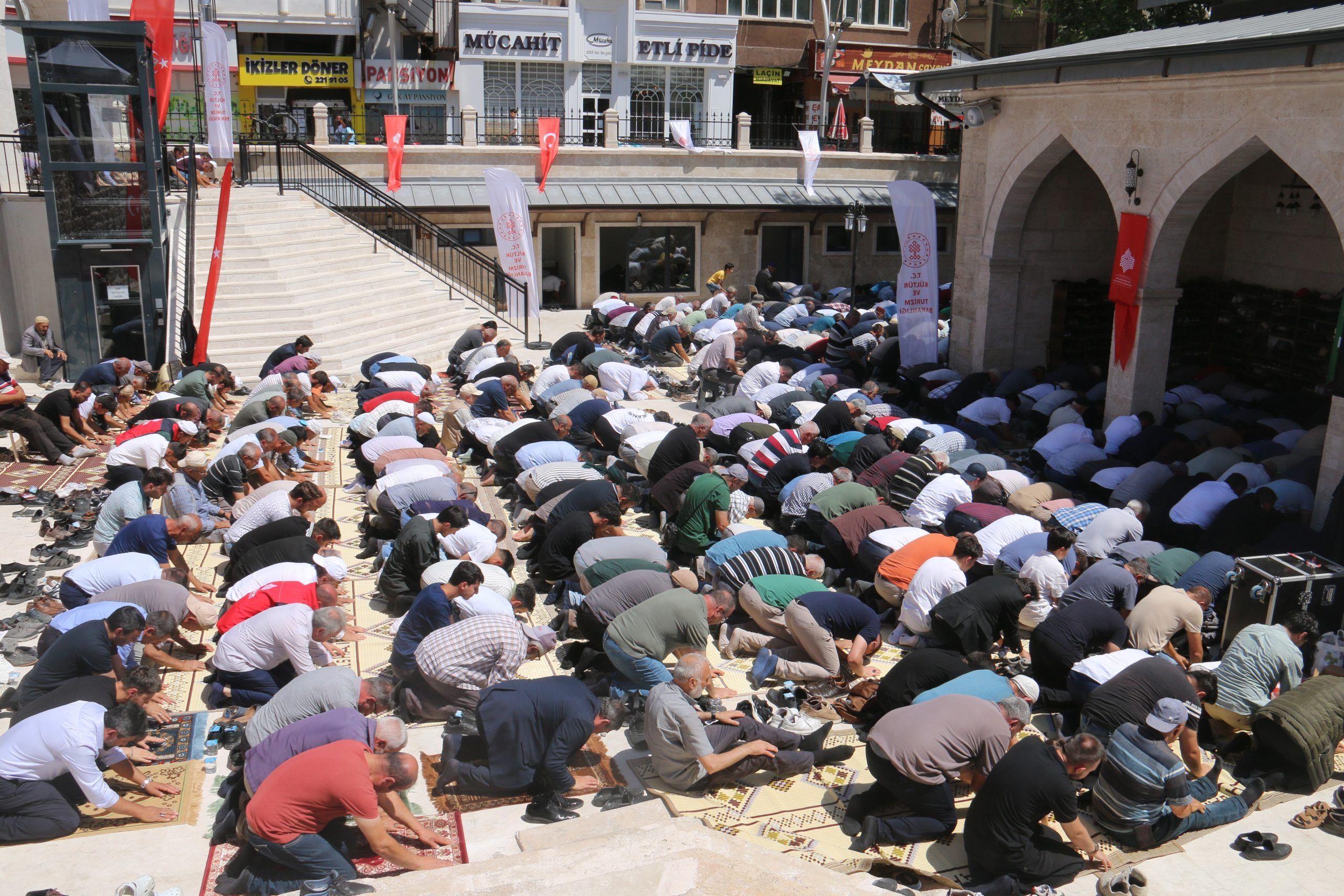 Cami avlusunda çok sayıda kişi saf hâlinde namaz kılarken, çevrede Türk bayrakları ve kemerli giriş görülüyor.