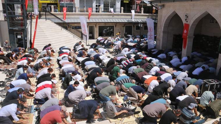 Cami avlusunda çok sayıda kişi saf hâlinde namaz kılarken, çevrede Türk bayrakları ve kemerli giriş görülüyor.