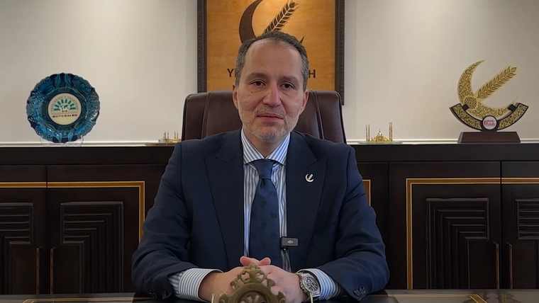 Bir masanın arkasında oturan bir kişi, kameraya doğru bakıyor ve gülümsüyor.