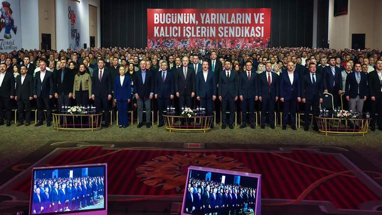 Bir salonda çok sayıda kişi sıralanarak duruyor; arkalarında “Bugünün, yarınların ve kalıcı işlerin sendikası” yazılı pankart var.