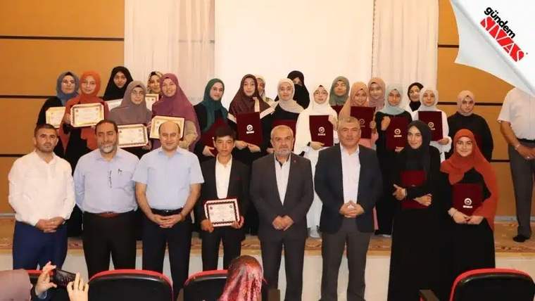 Bir salonda, ellerinde belgeler ve bordo kapaklı dosyalar bulunan kadın ve erkeklerden oluşan grup poz veriyor.