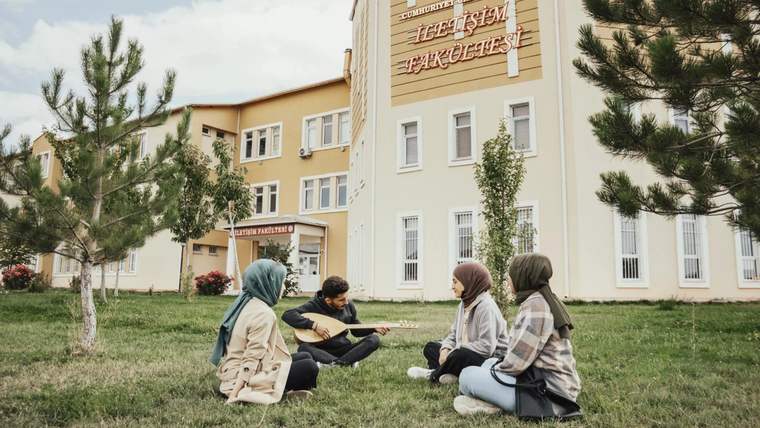 Sarı renkli eğitim binasının önündeki çim alanda dört genç oturuyor; çevrede çam ağaçları var.