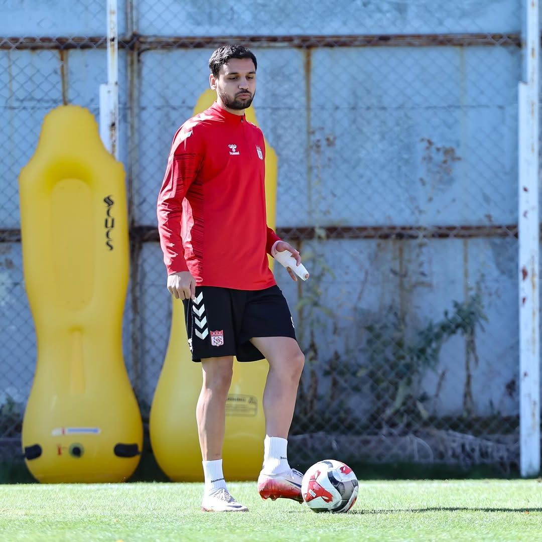 Kırmızı antrenman üstü ve siyah şort giyen bir kişi, sarı futbol antrenman mankenlerinin yanında yürüyor.