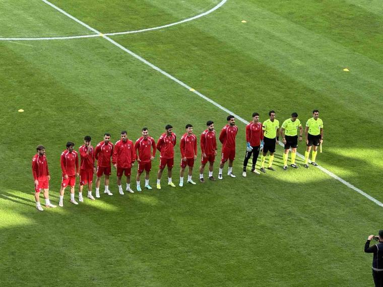 Futbol sahasında kırmızı formalı oyuncular, sarı formalı hakemlerin yanında yan yana duruyor.