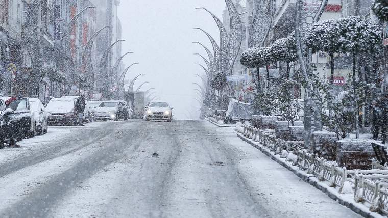 Kar yağışıyla kaplanan geniş caddede araçlar ilerlerken, kaldırımlar ve süs bitkileri beyaz örtüyle kaplanmış.