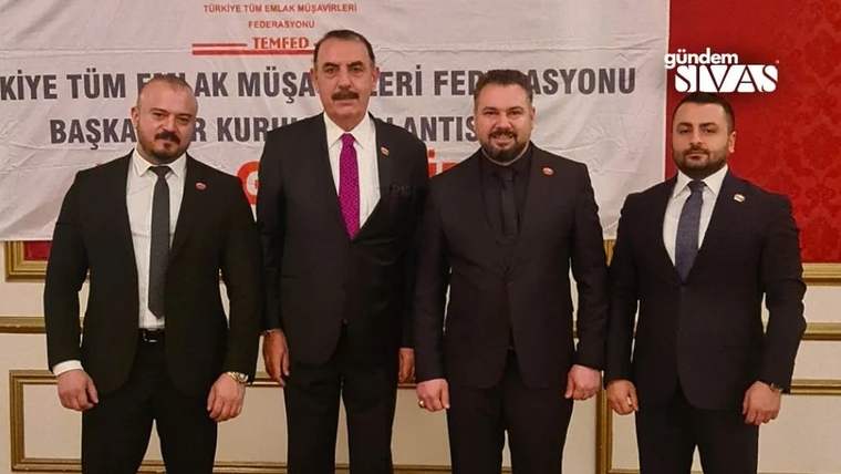 Dört kişi, arka planda bir pankartın önünde poz veriyor. Her biri takım elbise giymekte.