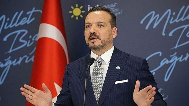 Mikrofon önünde konuşan takım elbiseli bir kişi, arkasındaki Türk bayrağı ve yazılı fonda el hareketi yapıyor.