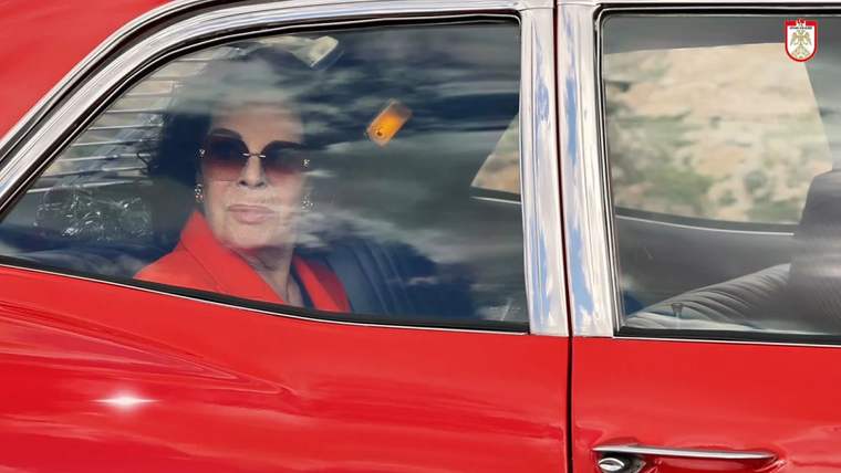 Kırmızı bir otomobilin arka koltuğunda oturan, güneş gözlüğü takan bir kadın görünüyor.