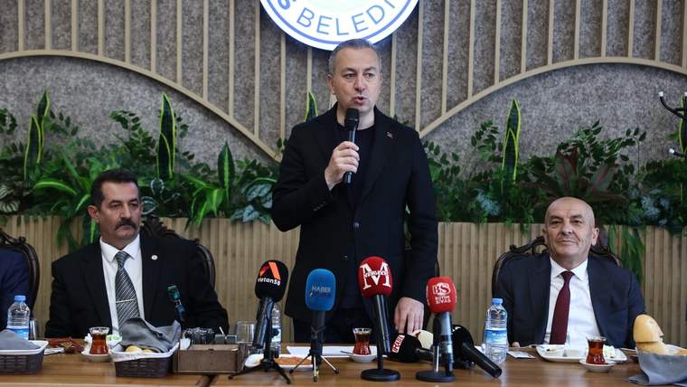 Masada çok sayıda mikrofon bulunan toplantıda, ortadaki kişi elindeki mikrofonla konuşurken iki kişi yanında oturuyor.