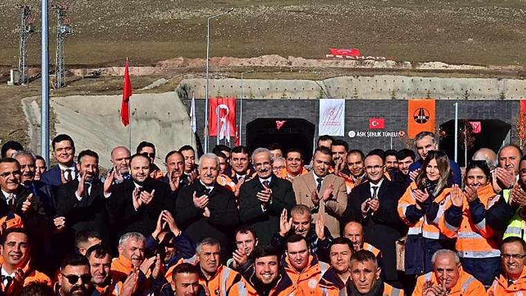 Kamu görevlileri ve işçiler, bir inşaat alanında alkışlayarak poz veriyor. Arkada Türk bayrağı ve çeşitli pankartlar görünmekte.