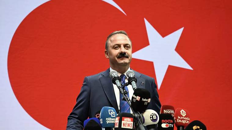 Mikrofonun önünde konuşan bir kişi, arkasında büyük bir Türk bayrağı var.