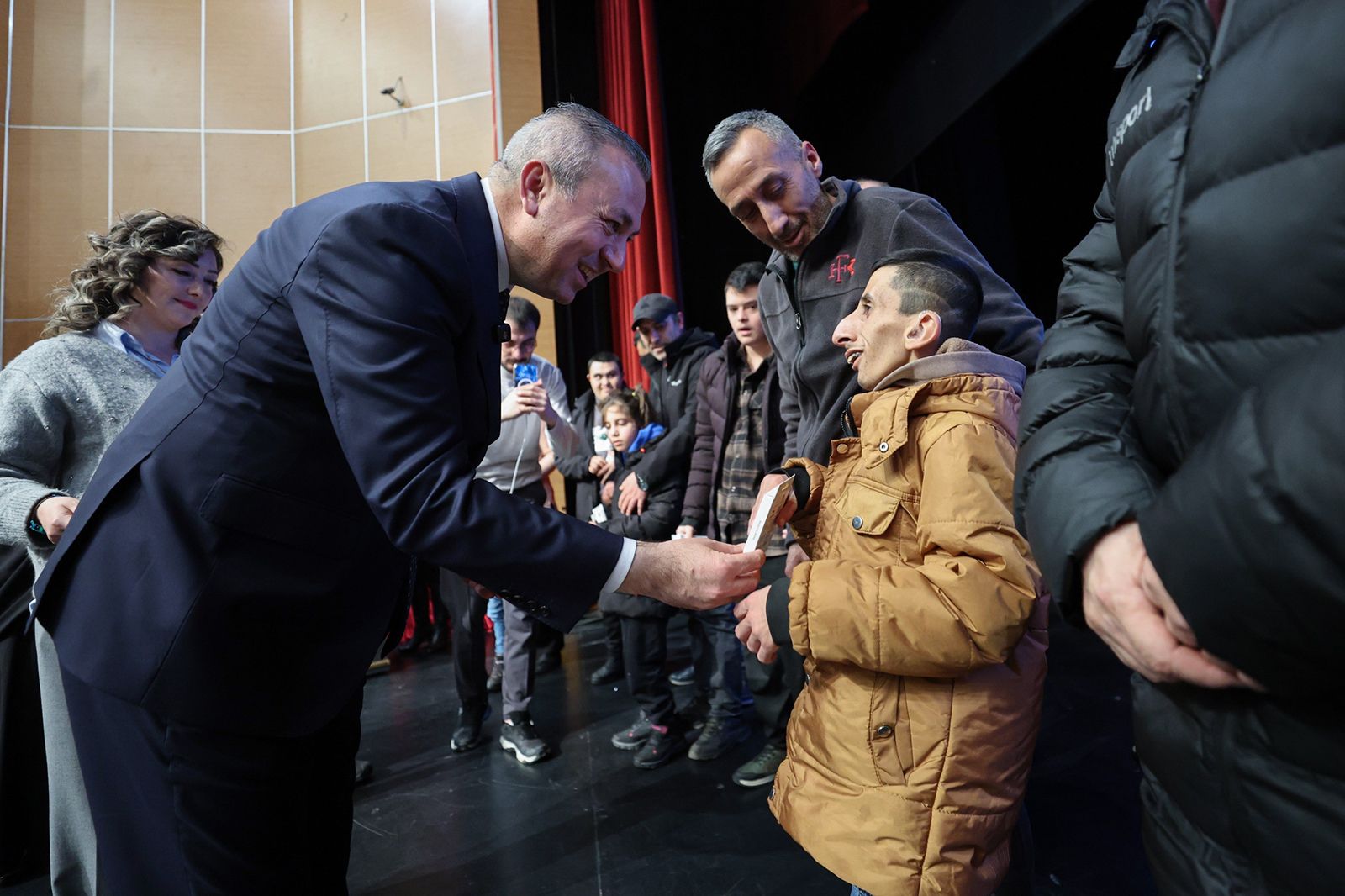 Bir kişi, sahnede bir çocuğa akıllı takip cihazı verirken gülümsüyor, etrafta başka insanlar var.