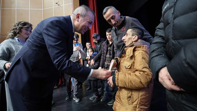 Bir kişi, sahnede bir çocuğa akıllı takip cihazı verirken gülümsüyor, etrafta başka insanlar var.