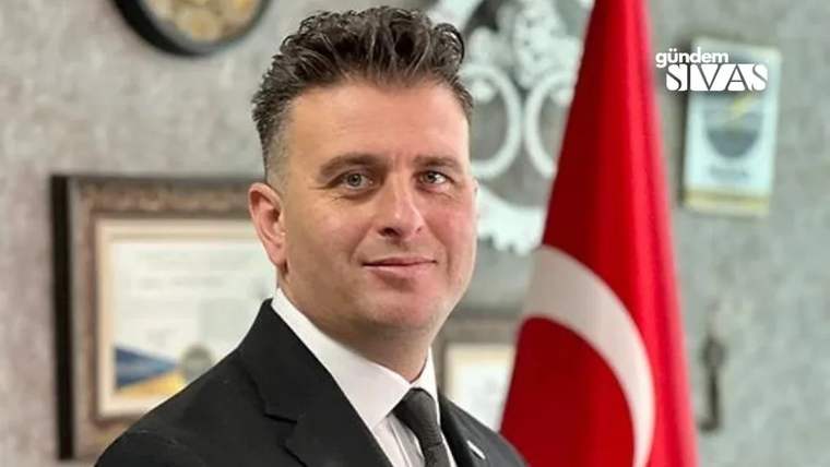 Kırmızı-beyaz Türk bayrağının önünde, takım elbise giymiş bir kişi gülümseyerek poz veriyor.