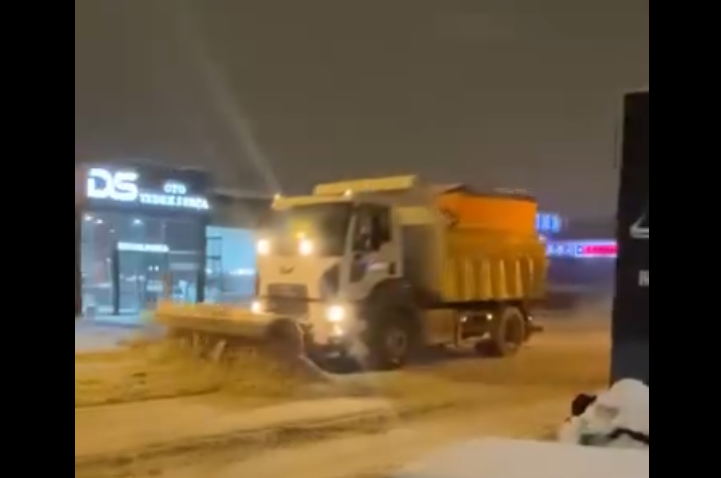 Karla mücadele aracı, karla kaplı bir sokakta tuz ve kum serpiştirerek ilerliyor.