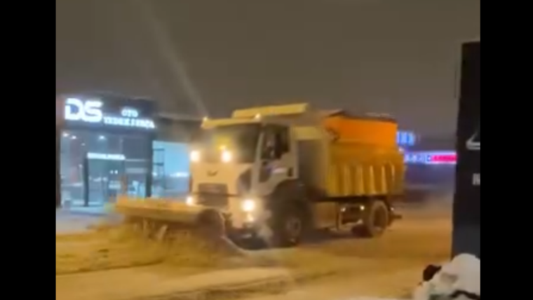 Karla mücadele aracı, karla kaplı bir sokakta tuz ve kum serpiştirerek ilerliyor.