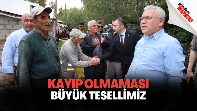 Açık havada bir grup erkekten biri yaşlı bir erkekle tokalaşırken, yanlarında takım elbiseli kişiler duruyor.
