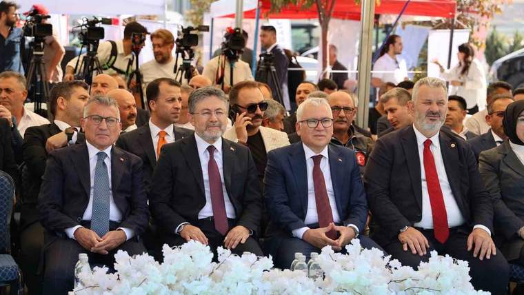 Ön sırada takım elbiseli dört kişi oturuyor; önlerinde beyaz çiçek aranjmanı, arkada kameralar görülüyor.