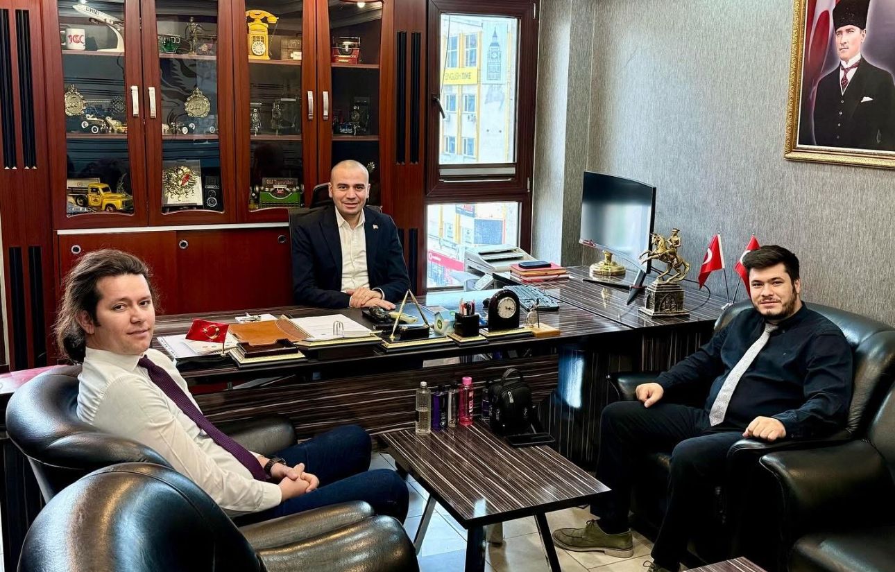 Bir masanın etrafında oturan üç kişi, arka planda Türk bayrağı ve Atatürk portresi var.