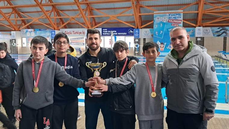 Kapalı spor salonunda dört öğrenci, bir yetişkinle birlikte kupa tutuyor; boyunlarında madalyalar bulunuyor.