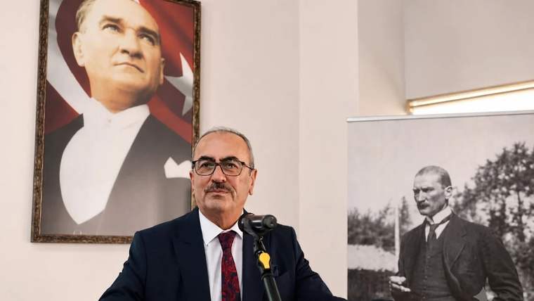 Kürsüde konuşan bir kişi, arkasında Atatürk portresi ve başka bir siyah-beyaz fotoğraf var.