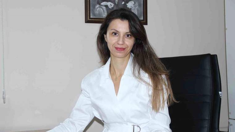 Beyaz önlüklü bir kadın, masa başında siyah koltukta otururken kameraya bakıyor.