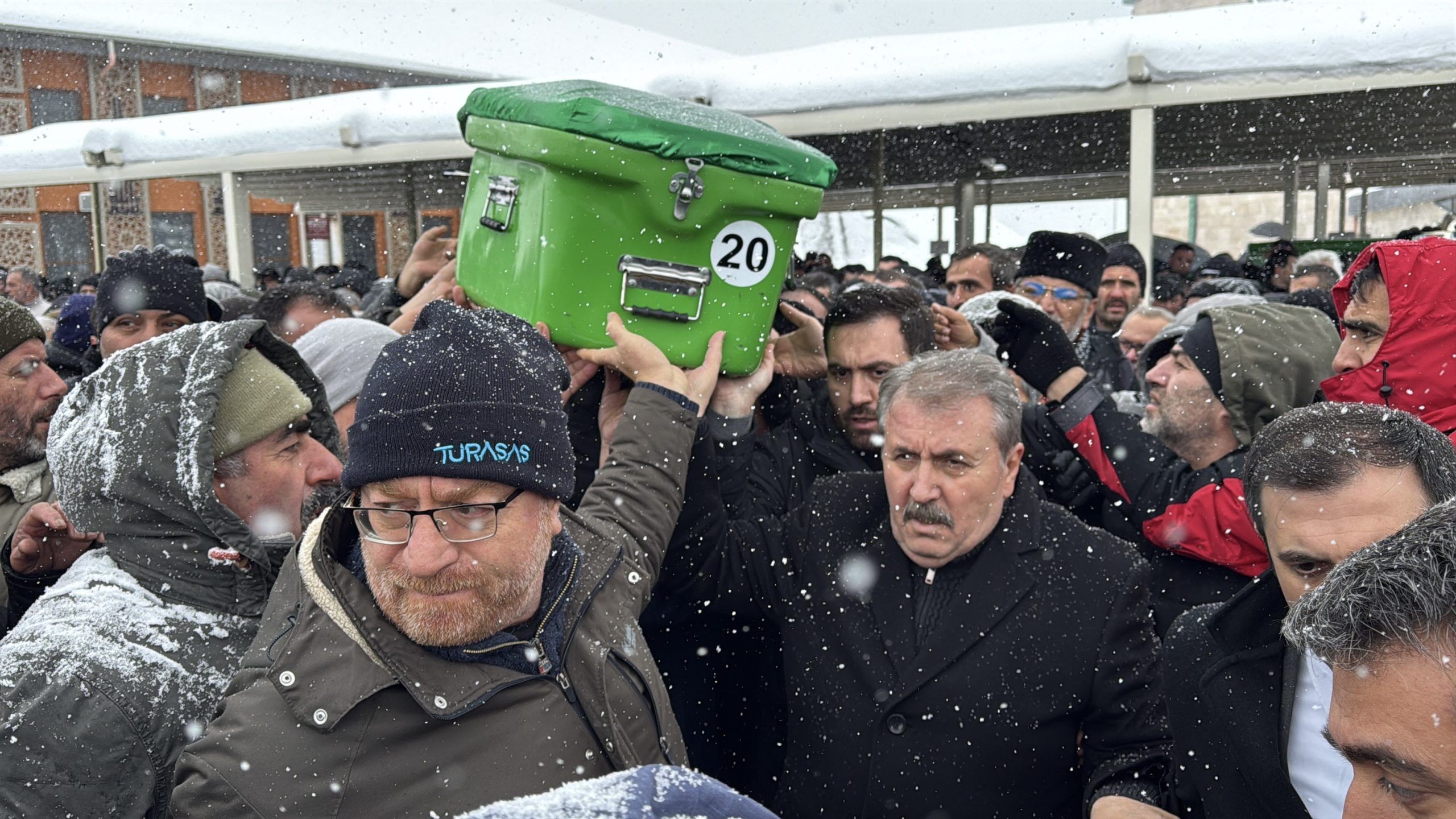 Kalabalık bir grup, yeşil bir tabutu taşırken kar yağışı altında duruyor.
