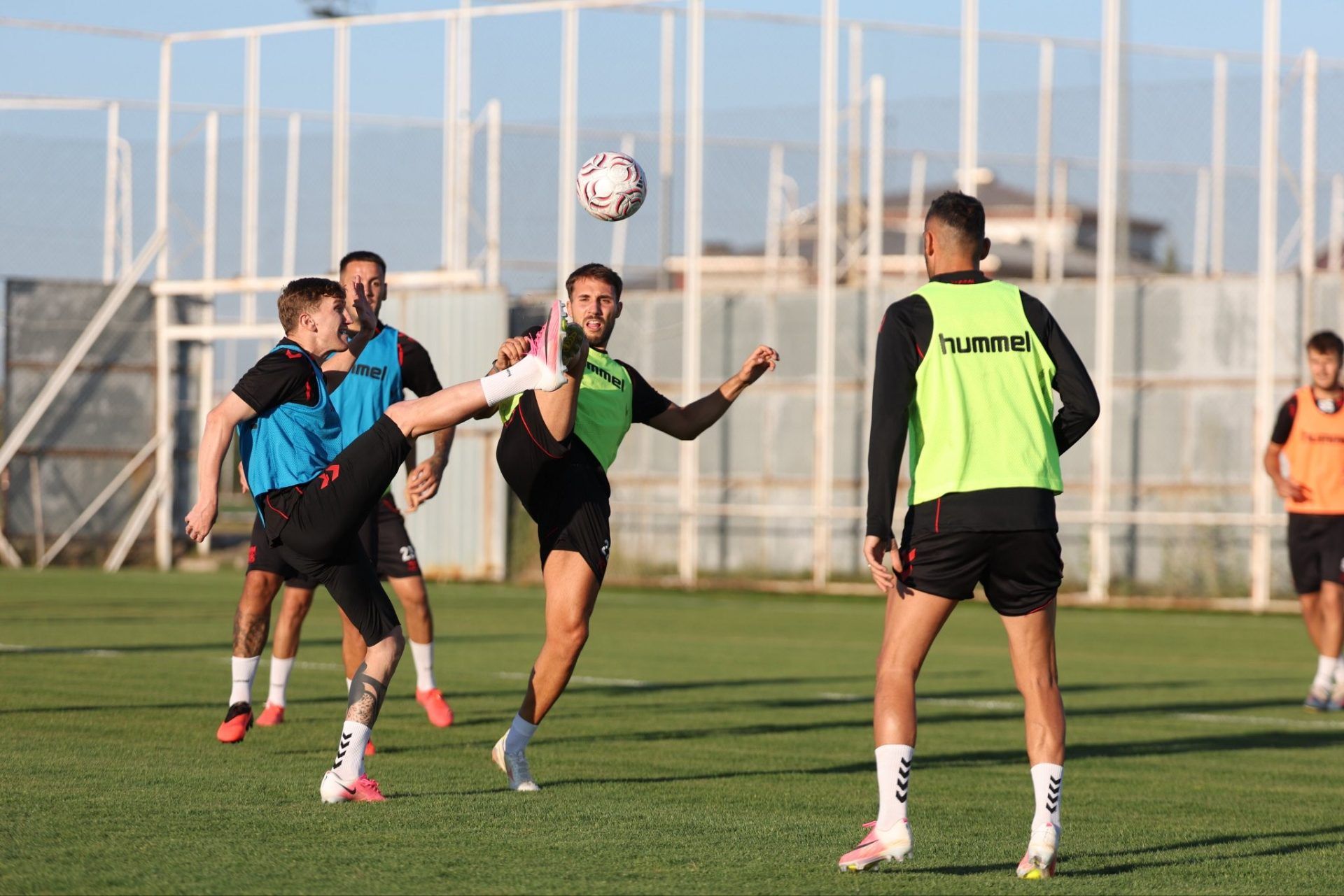 Yeşil sahada renkli antrenman yelekleri giyen futbolcular, havadaki top için mücadele ediyor.