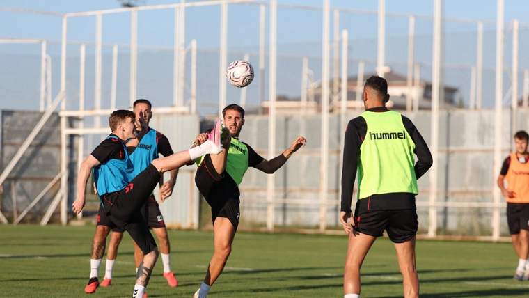 Yeşil sahada renkli antrenman yelekleri giyen futbolcular, havadaki top için mücadele ediyor.