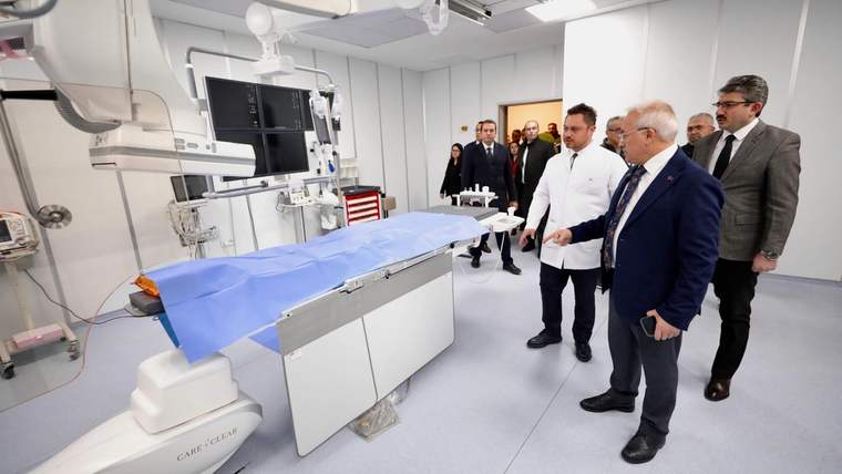 Anjiyografi cihazı ve üzeri mavi örtüyle kaplı hasta masası bulunan odada birkaç kişi duruyor.