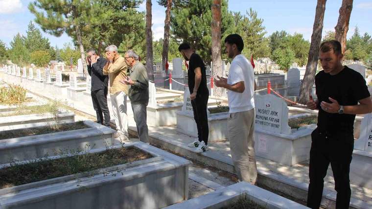 Çam ağaçları arasındaki mezarlıkta, mermer mezarların yanında duran birkaç kişi görülüyor.