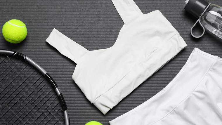 Beyaz bir tenis üstü, beyaz bir etek, iki yeşil tenis topu ve bir tenis raketi, su şişesiyle birlikte düz bir yüzeyde yer alıyor.