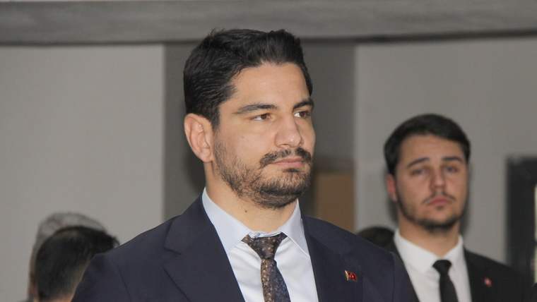 Takım elbiseli sakallı bir erkek önde dururken, arka planda kravatlı başka bir erkek görülüyor.