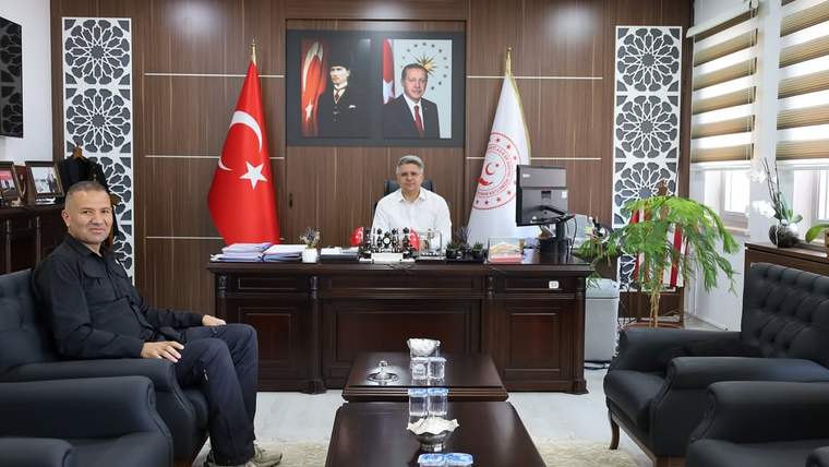 Makam odasında bir kişi masanın arkasında, diğeri koltukta oturuyor; Türk bayrağı ve flamalar görülüyor.