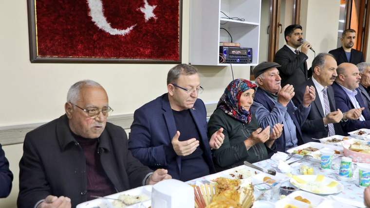 Duvarda ay yıldızlı Türk bayrağı motifi bulunan salonda, yemek masasındaki kişiler ellerini açmış; bir kişi konuşuyor.