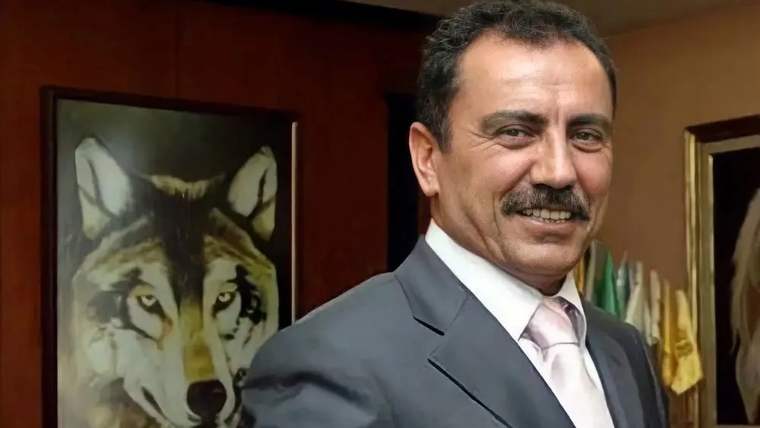 Gülümseyen bir adam, arkasında bir kurt resmiyle birlikte resmi bir kıyafet giymekte.
