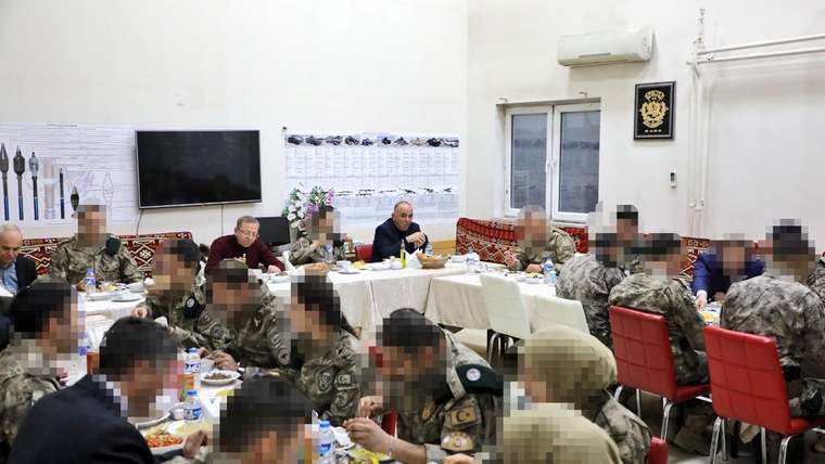 Kapalı bir salonda kamuflajlı ve sivil kıyafetli kişiler, masalarda yemek yerken görülüyor.