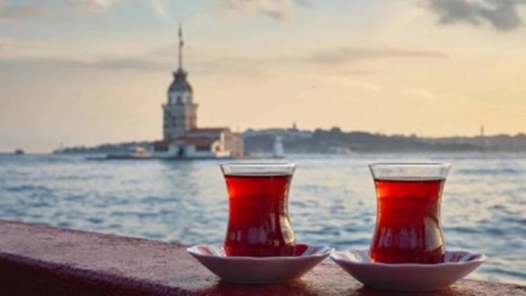 Deniz kıyısında korkuluk üzerindeki iki ince belli çay bardağı ve arkada kuleli yapı görülüyor.