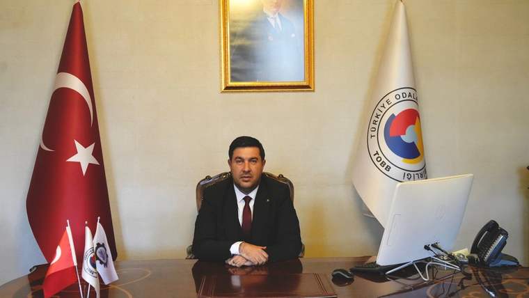Masanın arkasında oturan bir kişi, Türk bayrağı ve Türkiye Odalar ve Borsalar Birliği bayrağıyla görülüyor.