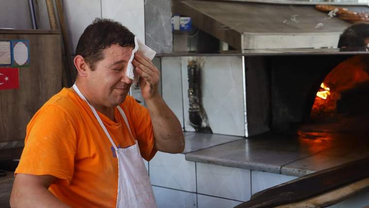 Turuncu tişörtlü ve beyaz önlüklü bir kişi, fırının yanında alnındaki teri mendille siliyor.