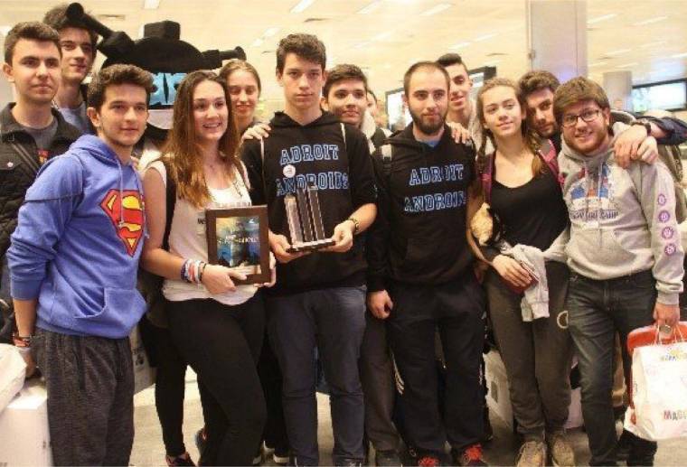 Kapalı bir mekânda bir araya gelen gençlerden biri plaket, diğeri kupa tutarken toplu poz veriyor.