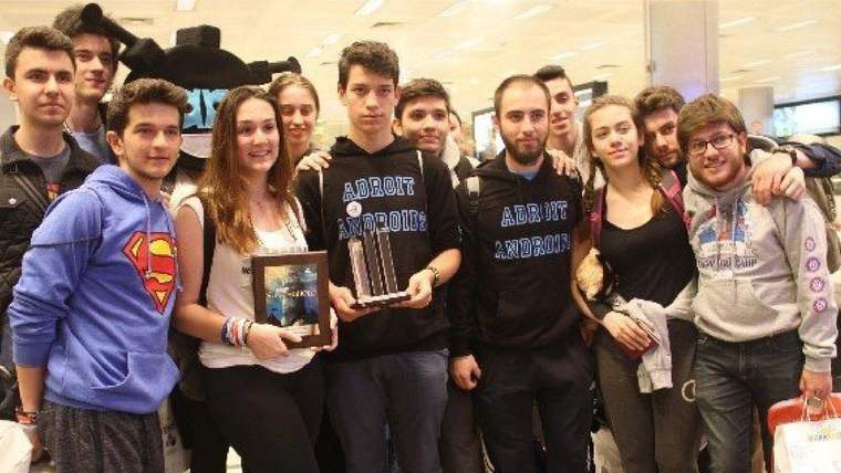 Kapalı bir mekânda bir araya gelen gençlerden biri plaket, diğeri kupa tutarken toplu poz veriyor.