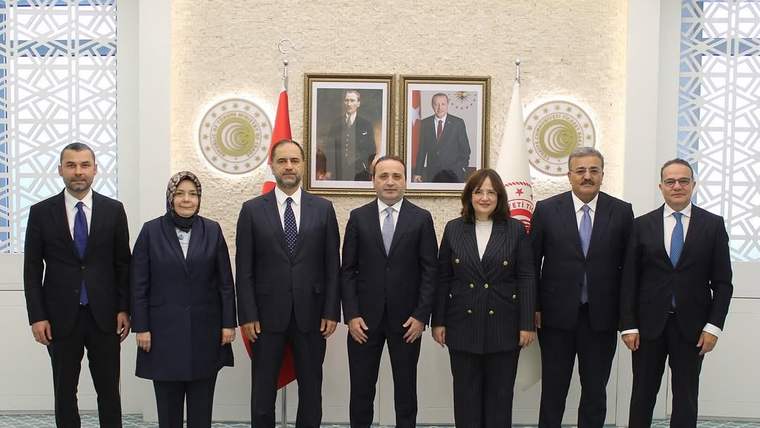 İç mekânda yedi kişi yan yana duruyor; arkalarında iki portre, bayraklar ve duvar süslemeleri görülüyor.